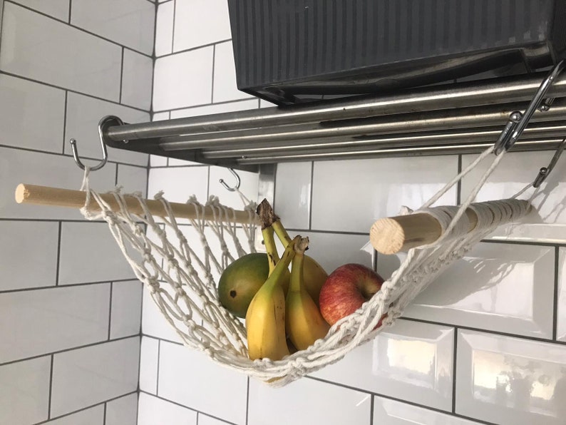 A white macrame hammock hanging from a metal shelf with a wooden dowel rod. The hammock is filled with yellow bananas and red apples.