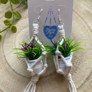 Puede incluir: Un par de pendientes de macramé con pequeñas macetas blancas y flores moradas. Los pendientes están hechos con cuerda de color crema natural y tienen ganchos plateados. Los pendientes están etiquetados como "Macra Made with Love".