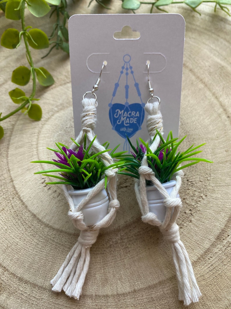 May include: A pair of macrame earrings with small white pots and purple flowers. The earrings are made with natural cream-colored string and have silver hooks. The earrings are labeled "Macra Made with Love".
