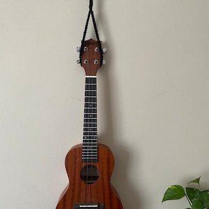 Może przedstawiać: Brązowy ukulele wiszący na haczyku na białej ścianie. Ukulele ma drewniane korpus i czarną główkę, a jest przymocowany czarną linką.