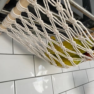 A white cotton rope hammock hanging from a metal shelf. The hammock is filled with green bananas and a red apple.