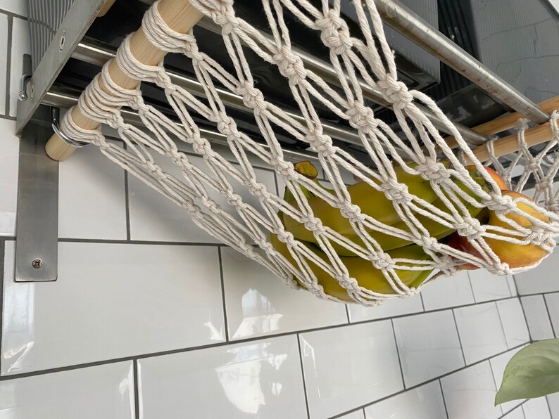 A white cotton rope hammock hanging from a metal shelf. The hammock is filled with green bananas and a red apple.
