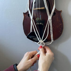 May include: A vintage violin with a macrame plant hanger. The violin is a dark brown color, and the macrame is white. The hands are holding the macrame. The background is a light gray wall.