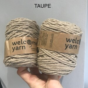 Close up of a hand holding two small skeins of taupe recycled cotton yarn against a grey painted wall