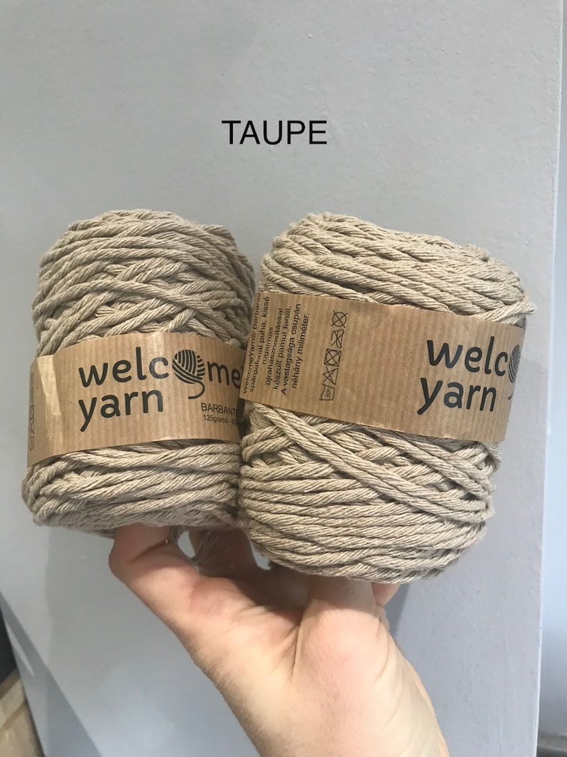 Close up of a hand holding two small skeins of taupe recycled cotton yarn against a grey painted wall