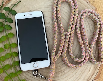 May include: A white smartphone with a black screen rests on a wooden surface, next to a pastel-colored macrame phone lanyard. The lanyard is made of woven cord in shades of pink, yellow, and green, and has a silver-colored clasp.