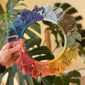 Puede incluir: Un tapiz de macram&eacute; de pared colorido con forma circular y detalles de flecos. El tapiz est&aacute; hecho con diferentes tonos de azul, verde, amarillo y naranja.