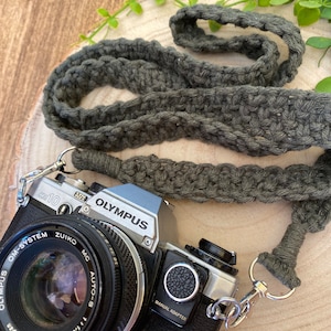 May include: A silver Olympus OM-10 camera with a black lens and a gray crocheted camera strap. The camera has a black manual adapter and the lens says "ZUIKO MC AUTO-S 1:1.8 f=50mm Japan".