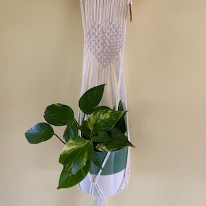 Könnte beinhalten: Ein weißer Makramee-Pflanzenhänger mit einem herzförmigen Design. Der Hänger besteht aus Baumwollschnur und hat oben einen Holzstab. Ein grün-weißer Topf mit einer grünen Pflanze hängt am Hänger.