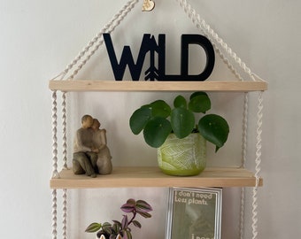 May include: Three wooden shelves hanging from a macrame rope with the word "WILD" in black letters on the top shelf. The shelves are decorated with plants, a small figurine, and a framed print that says "I don't need less plants, I need more shelves."