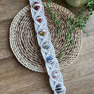 May include: A white macrame wall hanging with seven colourful stones and copper wire accents. The stones are arranged in a vertical line and are held in place by the macrame. The wall hanging is hanging from a wooden ring.