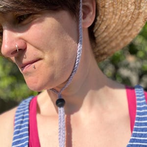 May include: A straw sun hat with a light purple chin strap and a black bead. The hat is worn with a blue and white striped tank top and a pink tank top underneath.