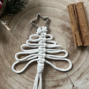 以下が含まれることがあります： 木製の表面に吊るされた、銀色の星がついた白いマクラメのクリスマスツリーのオーナメントです。