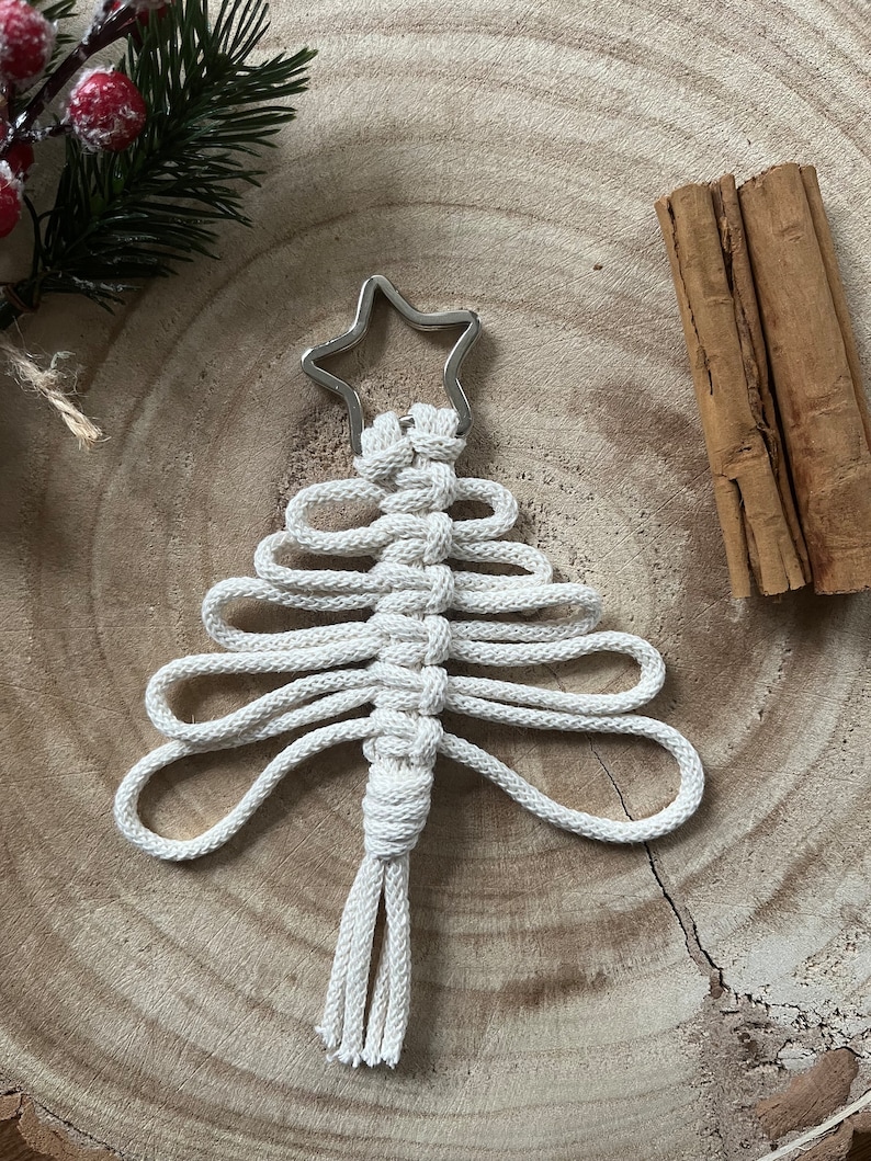 Puede incluir: Un adorno de &aacute;rbol de Navidad de macram&eacute; blanco con una estrella plateada en la parte superior. El adorno est&aacute; colgado en una superficie de madera.