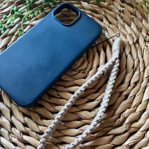 Può includere: Una custodia per telefono blu scuro con un cinturino per telefono intrecciato beige. La custodia del telefono e il cinturino sono su una superficie di paglia intrecciata.