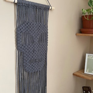 Puede incluir: Un tapiz de macramé azul grisáceo hecho a mano. La pieza de arte textil presenta un diseño geométrico y una barra de madera en la parte superior para colgarlo. El macramé tiene flecos en la parte inferior. Ideal para añadir textura y estilo.