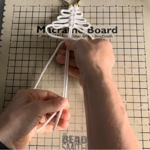 Puede incluir: Primer plano de las manos de una persona atando un nudo de macram&eacute; blanco en una tabla de macram&eacute; beige. La tabla tiene un patr&oacute;n de cuadr&iacute;cula y el texto "Macrame Board" est&aacute; impreso en la parte superior.