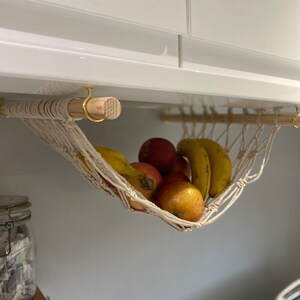 A white macrame hammock hanging from a white cabinet with a wooden dowel rod. The hammock is filled with yellow bananas and red apples.