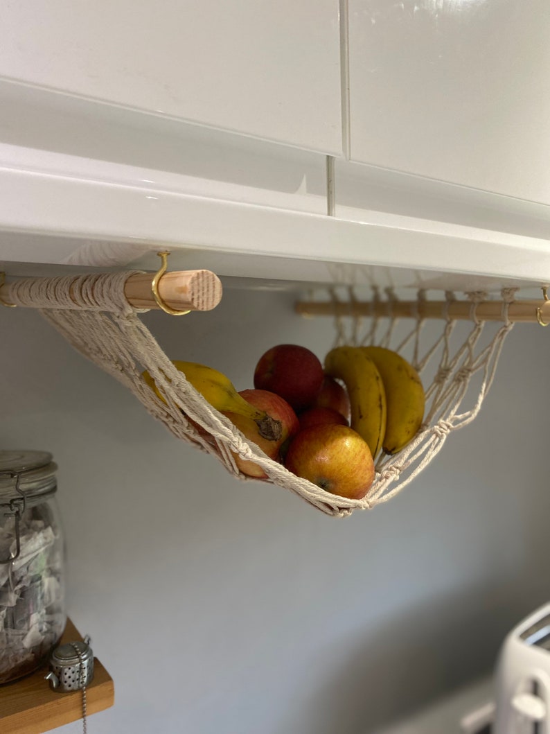 A white macrame hammock hanging from a white cabinet with a wooden dowel rod. The hammock is filled with yellow bananas and red apples.