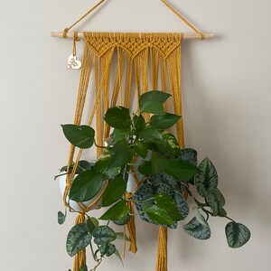 May include: A mustard yellow macrame plant hanger with two white pots and a green plant hanging from it. The hanger has a wooden dowel at the top and a small wooden heart tag that says "Home Sweet Home".