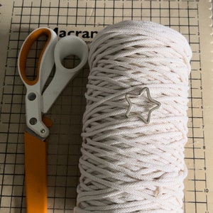 Puede incluir: Un rollo de cuerda de algod&oacute;n de color natural con un amuleto de estrella plateada, un par de tijeras naranja y blancas sobre una estera de corte cuadriculada.