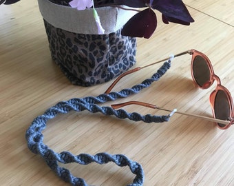 May include: A gray braided cord with plastic ends for holding glasses. The cord is looped around a pair of brown-tinted sunglasses. The sunglasses are lying on a wooden surface.