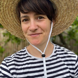 May include: A straw sun hat with a white chin strap is worn over a black and white striped shirt. The hat has a wide brim and a woven texture. The chin strap is adjustable with a black toggle. The shirt has horizontal stripes.