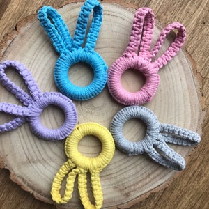 Op de afbeelding: Vijf gehaakte ringen in de vorm van een konijn in verschillende kleuren: lichtblauw, lavendel, geel, roze en grijs. De ringen zijn in een cirkel op een houten oppervlak gerangschikt.