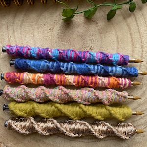 May include: Five colorful pens with a twisted yarn design. The pens are arranged vertically, with the top pen being pink, blue, and purple, followed by blue, orange, and pink, then pink, orange, and white, then green, and finally gold.