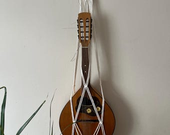 May include: A brown wooden mandolin suspended by a white macrame cord hanger. The hanger features intricate knotted patterns and a circular wooden ring at the top. The mandolin has a dark fretboard and a floral decal on the body near the sound hole.