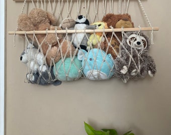 Puede incluir: Una hamaca de macramé beige con listones de madera cuelga de una pared, llena de animales de peluche, incluyendo un oso marrón, un pingüino, un perezoso y un lobo gris. La hamaca es una excelente manera de guardar y exhibir animales de peluche.