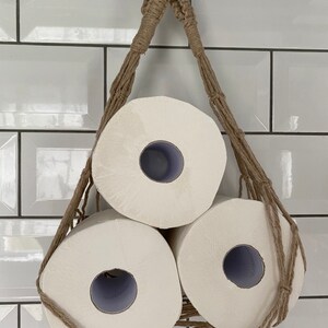 Front view of a handmade macrame toilet paper hammock, made with taupe recycled cotton, displayed with 3 large white toilet rolls inside, hanging against a white tiled wall