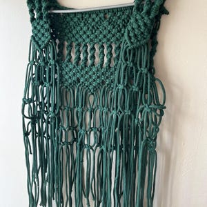 May include: A handmade, dark green macrame vest with a fringed bottom. The vest is hanging on a silver hanger against a neutral wall. The macrame design features a woven pattern with long, dangling fringe.