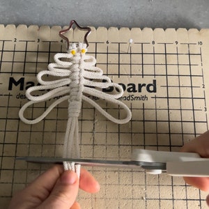 Puede incluir: Un adorno de &aacute;rbol de Navidad de macram&eacute; blanco con una estrella dorada en la parte superior. El adorno se est&aacute; recortando con unas tijeras sobre una estera de corte.