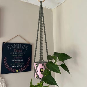 Peut inclure: Un porte-plante en macramé avec un pot rose et noir à imprimé léopard. Le porte-plante est suspendu à un anneau en bois et présente une plante verte. Un panneau mural indique "FAMILIES are like trees..."