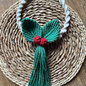 Può includere: Decorazione natalizia in macramè fatta a mano con un anello di corda bianca, foglie verdi, bacche rosse e un nappa verde. Il design festivo è ambientato su uno sfondo intrecciato, evocando un'estetica rustica delle feste.