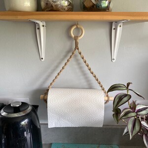 May include: A macrame paper towel holder with a wooden dowel and a roll of white paper towels. The holder is suspended from a wooden ring. The background includes a wooden shelf, white brackets, and a black kettle.