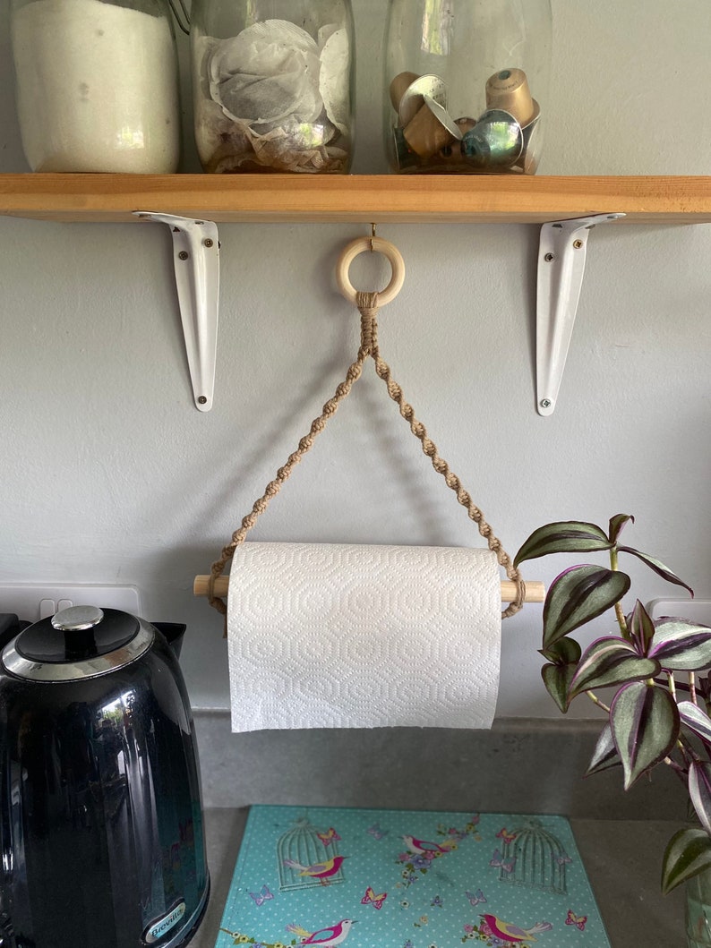 May include: A macrame paper towel holder with a wooden dowel and a roll of white paper towels. The holder is suspended from a wooden ring. The background includes a wooden shelf, white brackets, and a black kettle.