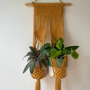 May include: A macrame plant hanger with two pots, one with a green plant and one with a purple plant. The hanger is made of mustard yellow cord and has a wooden dowel at the top.