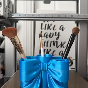May include: A bright blue 3D-printed cylindrical makeup brush holder featuring a prominent bow design on the front. The holder contains three makeup brushes with varying bristle colours and handle finishes, including gold, white marble, and black.
