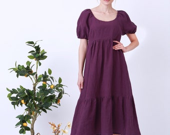 May include: A woman wearing a purple midi dress with puffed sleeves and a tiered skirt. She is standing in front of a white background with a potted plant and a vase of flowers.