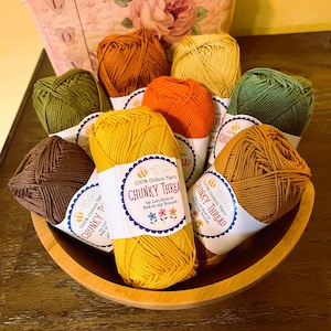 May include: A wooden bowl filled with colorful yarn skeins. The yarn is in shades of yellow, orange, brown, and green. Each skein has a label that reads "CHUNKY THREAD" and "100% Cotton Yarn".