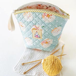 May include: A quilted, light blue yarn bag with a floral pattern and a small patch featuring a cartoon chicken. The bag is open, revealing a floral interior. A ball of yellow yarn, knitting needles, and a knitted swatch are also present.