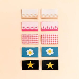 May include: Ten rectangular fabric labels arranged in two columns. The designs include two beige labels with white scalloped edges, two pink labels with white scalloped edges, two red and white gingham labels, two blue labels with white flowers, and two black labels with yellow stars.