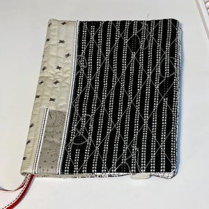 May include: A quilted book cover with a black and white striped pattern on the front and a beige fabric with spool designs on the spine. The cover has a red and white ribbon bookmark and a pocket with the text "Needles & Pins".