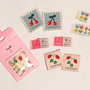 May include: A collection of woven labels with various designs. Includes labels with cherries, strawberries, and cross-stitch patterns. Some labels read "HANDMADE", "in EVERY stitch", and "HOMEMADE". A pink package contains additional labels.