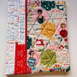 May include: A quilted fabric notebook cover with a patchwork design. The cover features a red zipper and a red fabric strip with white snowflake patterns. The patchwork includes hexagonal shapes with vintage illustrations and sewing-related text.