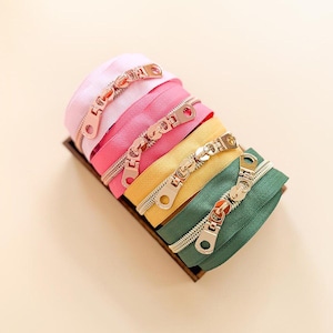 May include: A set of four coiled zippers in light pink, hot pink, yellow, and dark green, each featuring a metallic gold-tone pull tab. The zippers are arranged in a stack within a small rectangular wooden tray, showcasing their textured fabric.