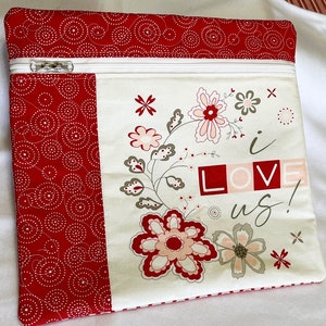 May include: A red and white zippered fabric pouch with a floral design and the words "i LOVE us!". The pouch has a red patterned border and a white zipper. The floral design includes red, pink, and gray flowers.