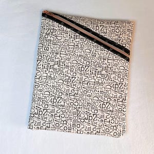 May include: A rectangular cream-colored zippered pouch with a black and white number and letter pattern. The zipper is black with a rose gold pull. The pouch is likely used for storing items.